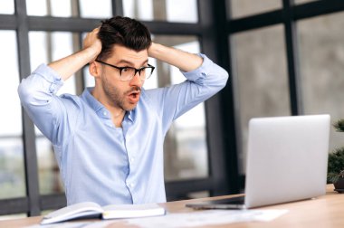 Shocked surprised caucasian man, businessman or office worker, sitting at his work desk, looking in amazement at the laptop, clutching his head with his hands, received unexpected news, won a victory