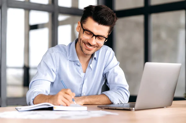 Hombre caucásico exitoso inteligente con gafas, sentado en el lugar de trabajo, usando una computadora portátil, tomando notas, estudiando o trabajando en línea, escuchando un webinar, ganando conocimiento, sonriendo —  Fotos de Stock