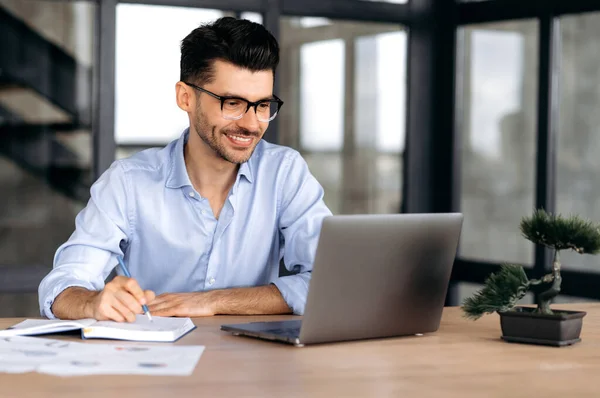 Online vzdělávání. Inteligentní úspěšný běloch s brýlemi, sedící na pracovišti, používající notebook, si dělá poznámky, studuje nebo pracuje online, poslouchá webináře, získává znalosti, usmívá se — Stock fotografie