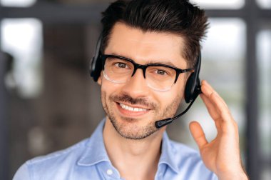 Close-up portrait of handsome satisfied man, operator of call center or business leader. Caucasian businessman with glasses, uses headphones for online conference, looks at camera and friendly smiling
