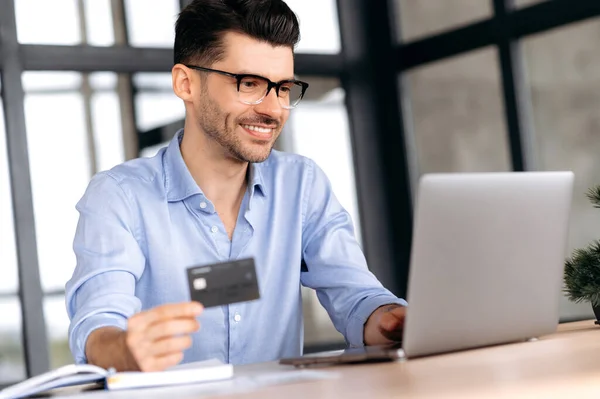 Online shopping, payment for purchases by credit card and laptop. Caucasian modern guy sitting at the table uses a laptop and a banking card to pay online purchases or delivery goods at home