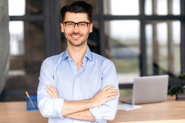 Formal business portrait. Confident successful caucasian businessman or manager with glasses stands near his work desk in the office, arms crossed, looks directly at the camera and smiles friendly