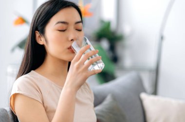 Sağlıklı bir yaşam tarzı. Güzel genç Asyalı kadın evdeki kanepede otururken bir bardak su içiyor. Güzel, mutlu, sağlıklı Japon kızı sağlıklı yaşam tarzını izler.