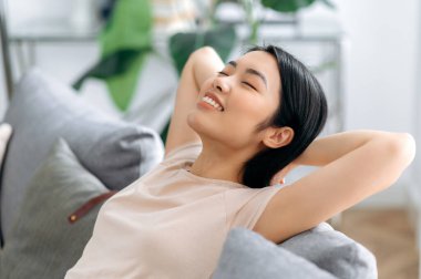 Tatmin olmuş Asyalı bir kadının oturma odasındaki kanepeye uzanmış gülümsemesi. Güzel Japon kızı evde takılıyor, tembellik ediyor, tatillerin hayalini kuruyor, ellerini başının arkasına atıyor.