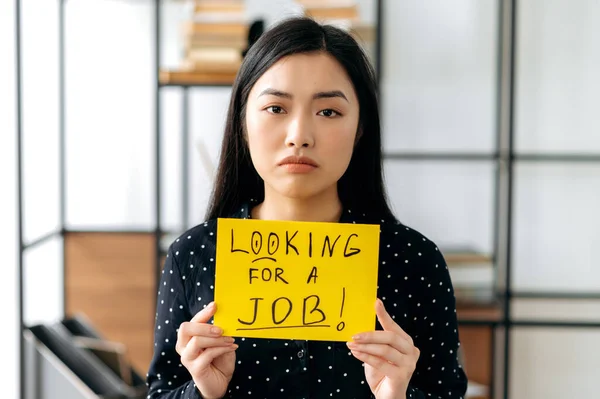 Portrait depressed unemployed young Asian woman, student or freelancer, sits at table with sign with inscription looking for a job, hopes to get job of her dreams, look sadly at camera — Stock Photo, Image