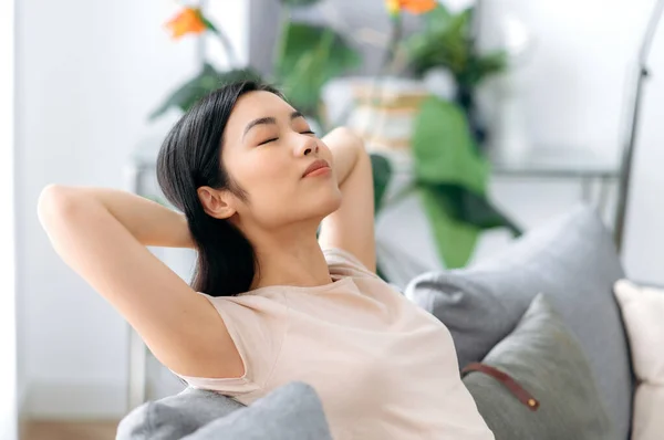 Calm beautiful young asian brunette woman, in casual t-shirt, relaxing on sofa in living room, resting at home, dreaming about vacation, throwing her hands behind her head, closing her eyes, chilling