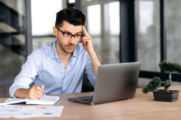 Enfocado pensativo joven gerente caucásico atractivo, freelancer o corredor con gafas sentados en la mesa en la oficina con estilo, utilizando el ordenador portátil, pasando por el entrenamiento en línea, lluvia de ideas, tomando notas — Foto de Stock