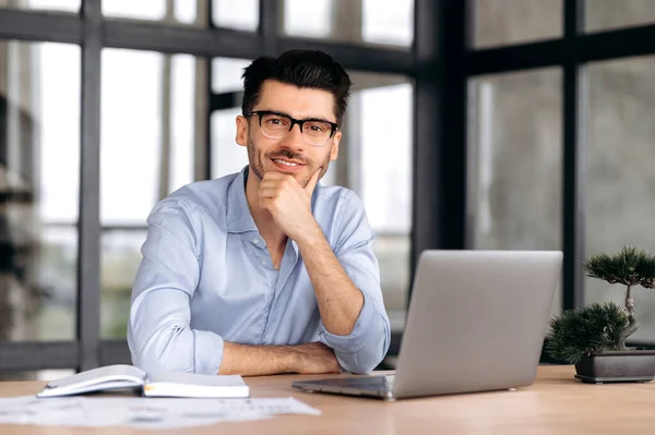 Portrait handsome young caucasian freelancer or manager, wearing strict shirt and glasses, sitting at table with laptop, supporting his head with his hand, looking at camera and smiling friendly