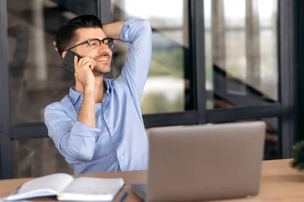 Sueño pacífico hombre caucásico con gafas, freelancer, gerente o corredor, sentado en el ordenador portátil en la oficina, inclinándose hacia atrás, hablando en el teléfono inteligente con amigos, tomando un descanso, mirando hacia otro lado, sonriendo — Foto de Stock