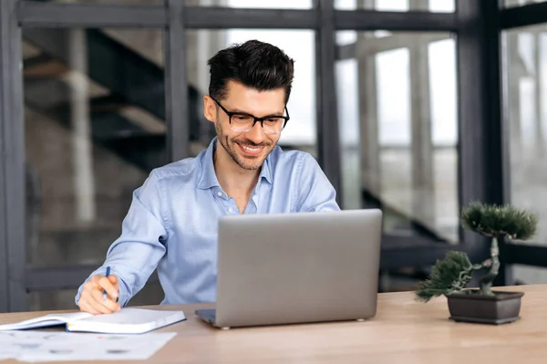 Joven hombre de negocios, corredor o gerente caucásico alegre y seguro en ropa formal y gafas sentados en la mesa en la oficina, tomando notas, usando el ordenador portátil, mirando a la pantalla, sonriendo —  Fotos de Stock