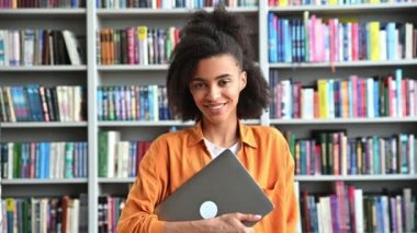 Öğrenci bir kızın video portresi. Afrikalı Amerikalı güzel, kendine güveni tam bir bayan öğrenci, şık giyinmiş, dizüstü bilgisayar tutuyor, bir üniversite kütüphanesinde duruyor, kameraya bakıyor, arkadaşça gülümsüyor.