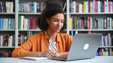 Çevrimiçi iletişim. Zeki, üniversite kütüphanesinde dizüstü bilgisayarla bir masada oturan, eğitici video dersleri alan, notlar alan, ekrana bakan Afro-Amerikan kız öğrencilere odaklanmış.