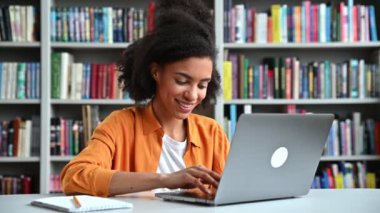 Kıvırcık saçlı, modaya uygun giyinmiş mutlu Afrikalı Amerikalı kız öğrenci, üniversite kütüphanesinde bir masada oturuyor, dizüstü bilgisayar kullanıyor, klavyeye yazı yazıyor, çevrimiçi derste notlar almaya odaklanıyor.