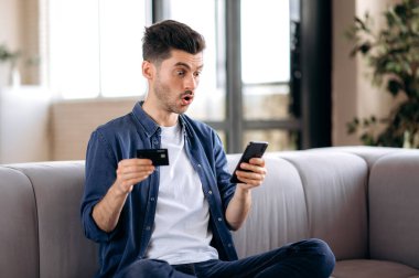 Şok olmuş çekici beyaz bir adam oturma odasındaki koltukta oturuyor, cep telefonu ve banka kartını internet üzerinden alışveriş ve internet üzerinden alışveriş yapmak için kullanıyor.