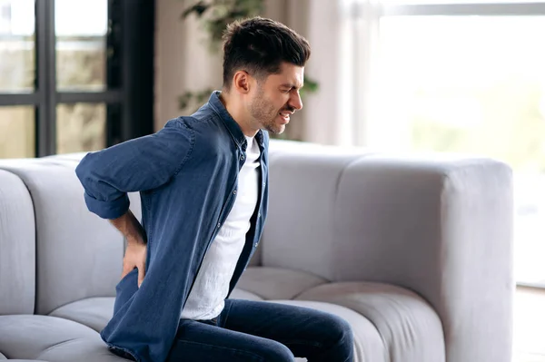 Triste joven adulto caucásico barbudo hombre, en ropa casual, sentado en el sofá en la sala de estar, masajeando su espalda, sufriendo de dolor de espalda, sufriendo de artritis, ciática —  Fotos de Stock