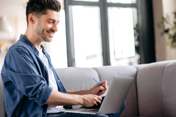 Smart självsäker ung kaukasisk man sitter på soffan, i casual kläder, student gör online distansundervisning, frilansare arbetar distans, anteckna, titta på skärmen, ler — Stockfoto