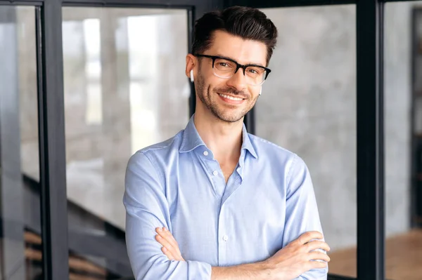 Portrait of joyful attractive confident young adult business man or manager wearing glasses and headphones, wearing formal shirt, standing in office, looking at camera, arms crossed, smiling friendly — Stock Photo, Image