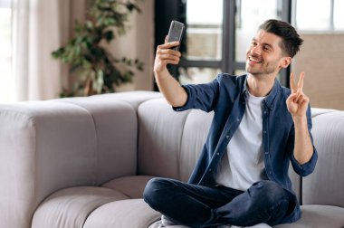 Mutlu, yakışıklı beyaz adam, günlük kıyafetler içinde, dinleniyor, lotus pozisyonunda koltukta oturuyor, elinde bir cep telefonu tutuyor, selfie çekiyor, eliyle işaret ediyor, gülümsüyor.