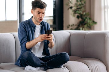Kafası karışmış genç beyaz bir adam, günlük kıyafetleriyle, Lotus pozisyonunda koltukta oturuyor, akıllı telefon kullanıyor, beklenmedik bir mesaj alıyor, arkadaşlarıyla mesajlaşıyor, ekranda şaşkınlıkla bakıyor.