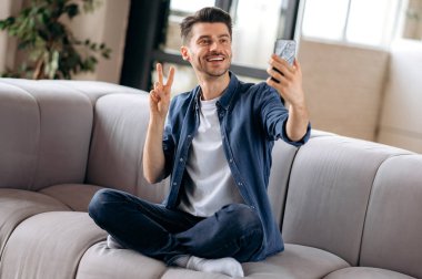 Çekici pozitif beyaz adam, şık kıyafetler içinde, dinleniyor, bir koltukta oturuyor, elinde bir akıllı telefon tutuyor, bir selfie çekiyor, eliyle işaret ediyor, arkadaşlarıyla görüntülü konuşma yapıyor, gülümsüyor.