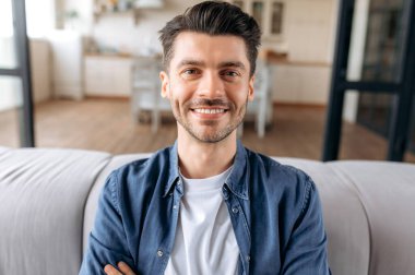 Yakışıklı, neşeli, beyaz bir adamın yakın plan portresi. Kollarını kanepede kavuşturmuş, şık bir kot gömlek giymiş, kameraya bakıyor ve dostça gülümsüyor.