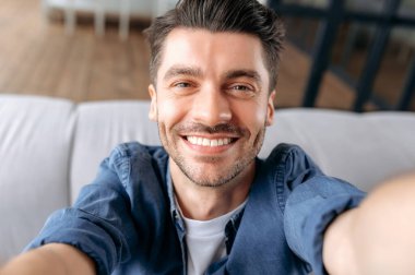 Genç, beyaz bir adam selfie çekiyor. Çekici, neşeli bir adamın portresi, kot gömlekli, koltukta oturuyor, akıllı telefonu elinde tutuyor ve webcam 'e bakıyor, arkadaşça gülümsüyor.