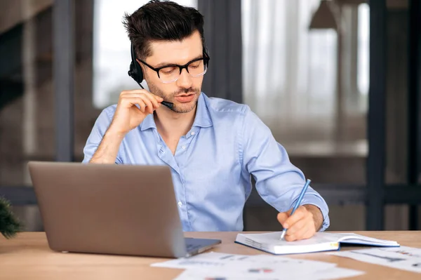 Fokuserad smart framgångsrik ung kaukasisk affärsman, frilansare eller konsult bär glasögon och hörlurar, sitter på kontoret, använder laptop, chatta med klienten online, anteckna — Stockfoto