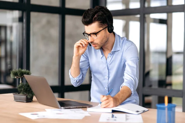 Homem caucasiano concentrado com óculos e fones de ouvido, funcionário de escritório ou consultor, fica perto do local de trabalho no escritório, se comunica com colegas ou cliente em uma conferência on-line, toma notas — Fotografia de Stock