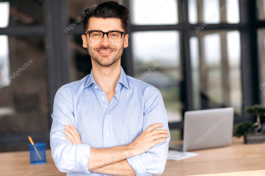 Attractive confident successful caucasian businessman or manager wearing glasses stands near his work desk in the office, in formal clothes, arms crossed, looks directly into camera, smiles friendly