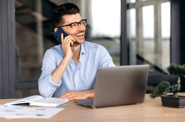 Mobile phone conversation. Happy caucasian freelancer or manager talking on a smartphone while working on a laptop, a positive man makes a pleasant phone conversation, looks away, smiles friendly