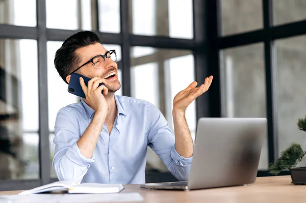 Comunicación con el teléfono celular. Amistosa persona de negocios caucásica alegre se sienta en la mesa en la oficina, se comunica emocionalmente por teléfono inteligente con amigos o colegas, gestos con la mano, mira hacia otro lado, sonríe —  Fotos de Stock