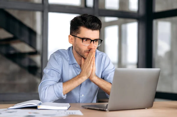 Hombre de negocios caucásico guapo concentrado, corredor o gerente con gafas, sentado en la mesa en la oficina, con la esperanza de éxito en el trabajo, mirando con esperanza a la pantalla, las manos se unen —  Fotos de Stock