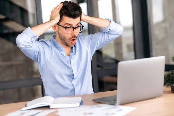 Shocked caucásico gerente masculino, corredor o freelancer, trabajando en la oficina, utilizando el ordenador portátil, incurriendo en enormes pérdidas, sobrecargado de trabajo, estresado, mirando la pantalla en sorpresa, sosteniendo la cabeza con las manos — Foto de Stock