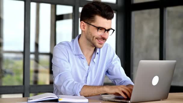 Online education. Intelligent successful caucasian man with glasses, sitting at the workplace, using a laptop, takes notes, studying or working online, listening to a webinar, gaining knowledge, smile — Stock Video