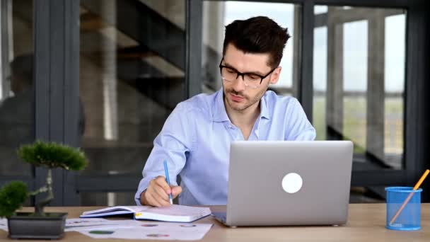 Smart framgångsrik kaukasisk man med glasögon, sitter på arbetsplatsen, använder en bärbar dator, antecknar, studerar eller arbetar online, lyssnar på ett webinar, får kunskap, ler — Stockvideo