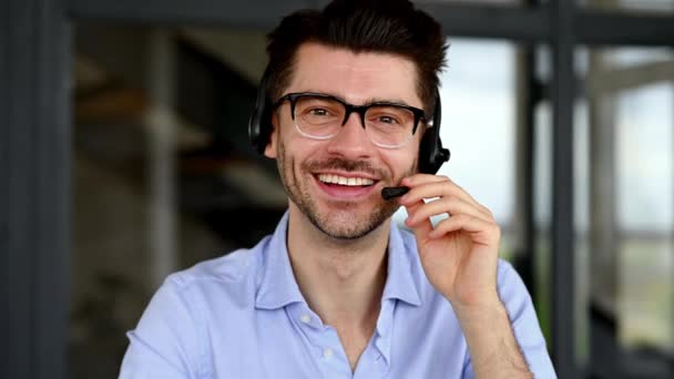 Hombre alegre guapo satisfecho, operador de centro de llamadas o líder de negocios con auriculares. Hombre de negocios caucásico con gafas, charlas a través de videoconferencia en línea, mira a la cámara y sonrisas amistosas — Vídeos de Stock