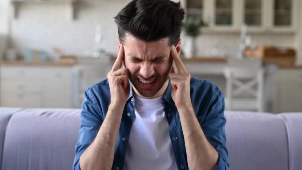 Headache, migraine. Caucasian young adult guy sitting at home on the sofa in the living room, massaging temples, suffering from headache, migraine, stress — Stock Video