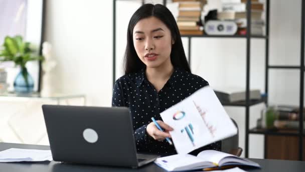 Amichevole donna d'affari asiatica sicura o insegnante in camicia formale, seduta alla sua scrivania, analizzando il rapporto, mostrando grafico finanziario ai dipendenti durante la videocomunicazione durante la chiamata in conferenza, sorridente — Video Stock