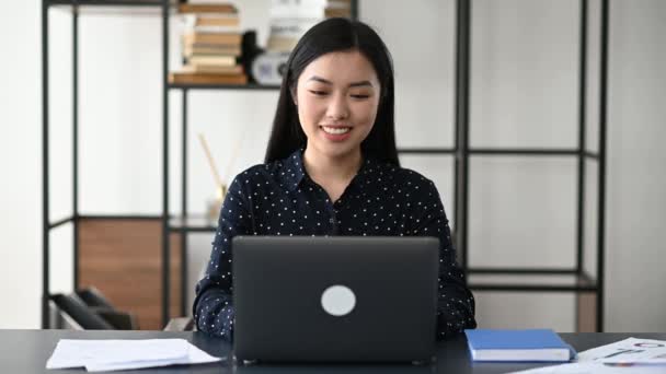 Vänlig söt säker ung asiatisk kvinna frilansare, chef, VD, sitter på kontoret på laptop, talar genom videosamtal med kunden eller anställd, diskutera affärsstrategi, viftande hand, leende — Stockvideo