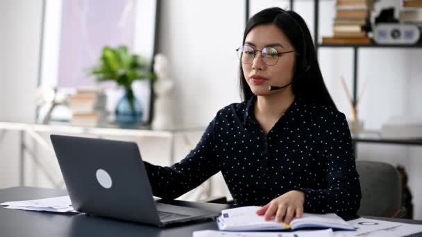 Pewna siebie, mądra azjatycka dziewczyna w okularach i słuchawkach, freelancer, operator call center, siedzi przy biurku w biurze, rozmawia z klientem lub pracownikiem przez komunikację wideo, gestykulując ręką — Wideo stockowe