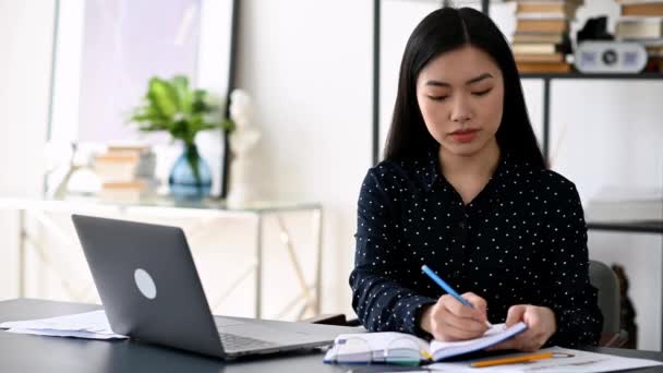 Cute inteligentny pewny azjatycki dziewczyna, freelancer lub student siedzi w pracy biurko, przy użyciu laptopa do pracy lub nauki, notatki podczas webinarium online lub kształcenia na odległość, patrząc na ekranie, uśmiechnięty — Wideo stockowe