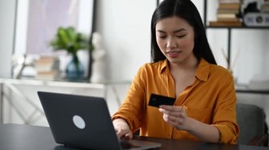İnternetten alışveriş. Masada oturan güzel Asyalı esmer kadın bilgisayarını ve kredi kartını online alımları ve teslimatları karşılamak için kullanıyor, veri giriyor, karta bakıyor, gülümsüyor.