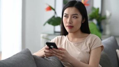 Neşeli Japon esmer kız, akıllı telefonunu günlük tişörtle koltukta otururken kullanıyor, internette ve sosyal ağlarda geziniyor, arkadaşlarıyla mesajlaşıyor, iyi haberleri alıyor, gülümsüyor.