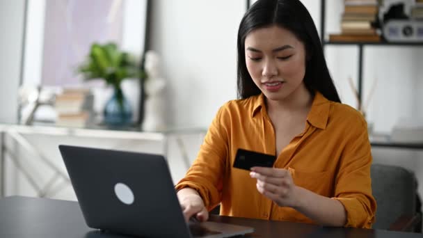 İnternetten alışveriş. Masada oturan güzel Asyalı esmer kadın bilgisayarını ve kredi kartını online alımları ve teslimatları karşılamak için kullanıyor, veri giriyor, karta bakıyor, gülümsüyor. — Stok video