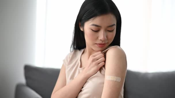 Feliz hermosa mujer joven asiática, vestida con camiseta, con un yeso adhesivo en el hombro después de ser vacunada de COVID 19 o gripe, mirando a la cámara, sonríe con gusto — Vídeos de Stock