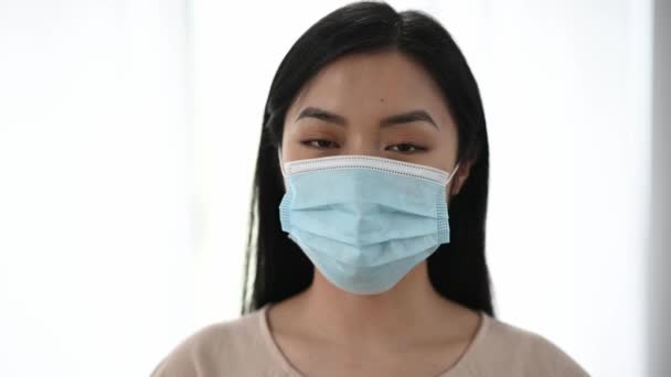 Medidas de prevenção, conceito de saúde. Close up de asiático chinês mulher no casual desgaste de pé dentro de casa com proteção médica máscara no seu rosto e olhando diretamente para a câmera — Vídeo de Stock