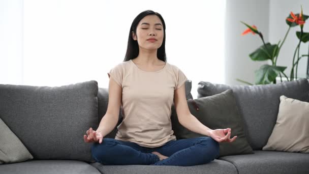 Ruhige, schöne junge asiatische Brünette, in lässiger Kleidung, zu Hause auf dem Sofa in Lotusposition sitzend, sich ausruhend, sich um ihre Gesundheit kümmernd, entspannend und meditierend, die Augen schließend — Stockvideo