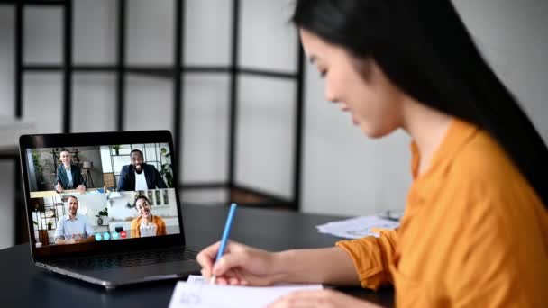 Videollamada, conferencia en línea con aplicación y portátil. Mujer asiática satisfecha estudiante o freelancer, estudia o trabaja a distancia, ve una conferencia en línea, toma notas, personas multirraciales en la pantalla del ordenador portátil — Vídeos de Stock