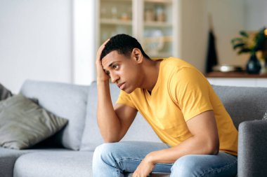 Üzgün, yakışıklı, yalnız, genç, İspanyol bir adam, evde, koltukta oturuyor, alnını tutuyor, baş ağrısı, depresyon, hayal kırıklığı, psikolojik desteğe ihtiyacı var.