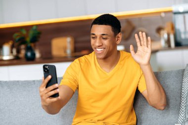 Akıllı telefon kullanarak video konferansında sohbet. Pozitif ispanyol bir adam oturma odasında bir kanepeye oturur, bir akıllı telefon tutar, video görüşmesiyle konuşur, bir el hareketiyle müfettişi selamlar, gülümser.